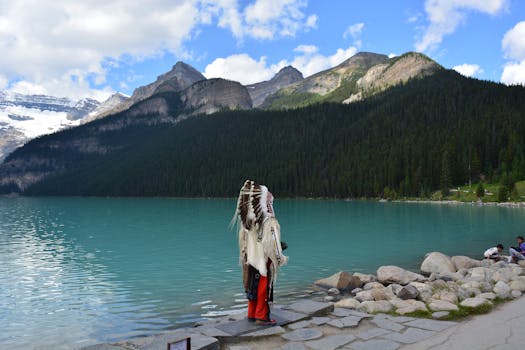Indigenous man in traditional headwear stands by Lake Louise with stunning mountain backdrop.